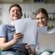 Happy couple reviewing structured settlement documents on couch while using laptop for financial planning