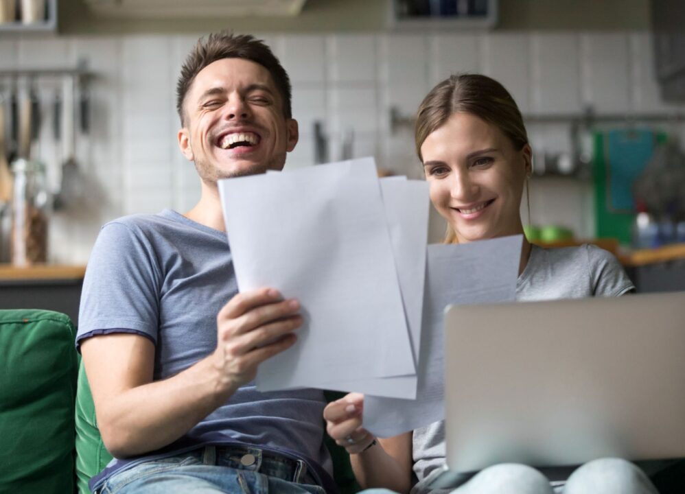 Happy couple reviewing structured settlement documents on couch while using laptop for financial planning