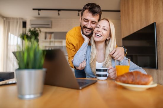 Happy couple sitting together at a table, smiling while looking at a laptop preparing a wellness budget.